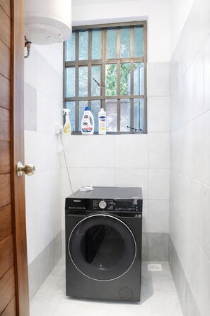Een kleine wasruimte met een voorlader wasmachine, een raam met tralies en een klein plankje met schoonmaakmiddelen.