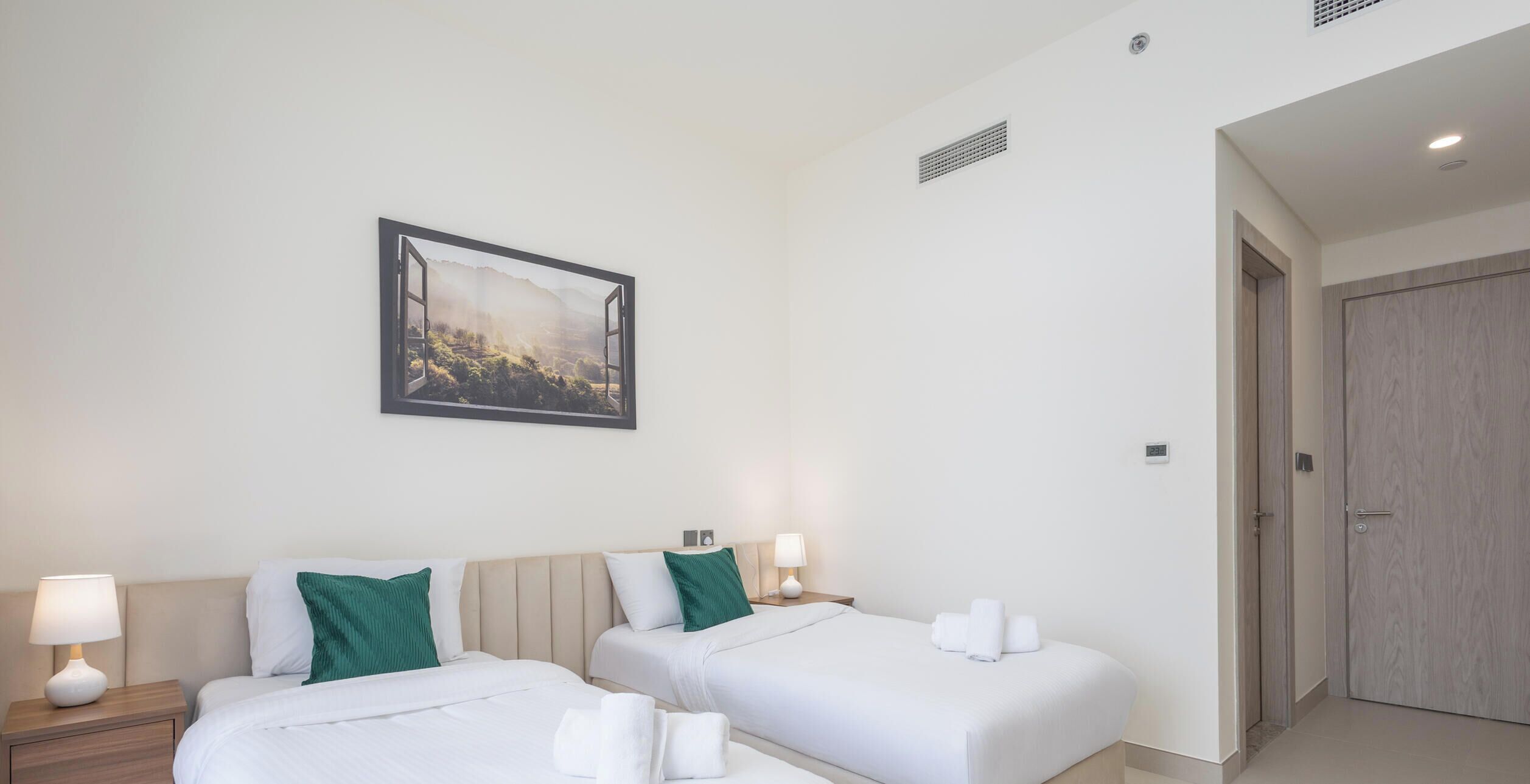 A hotel room with two beds, a painting of a landscape, a door, and a nightstand with a lamp.