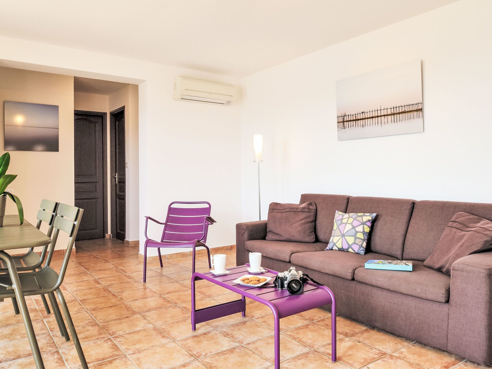 Una sala de estar con un sofá marrón, una silla morada, una mesa de comedor con sillas y una mesa baja morada con una cámara y un libro.