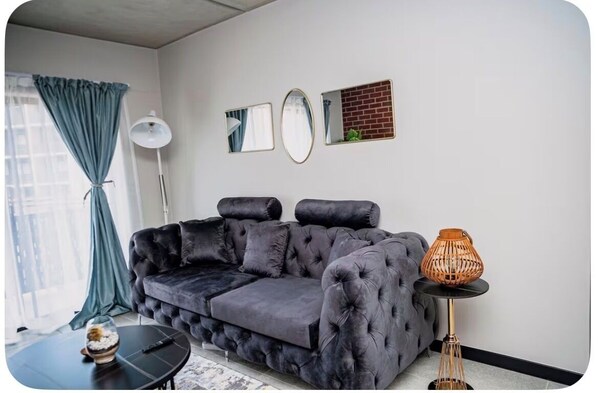 Een moderne woonkamer met een donkergrijze gestuufde bank, een ronde zwarte salontafel, een vloerlamp en een kleine tafel met een geweven lamp.