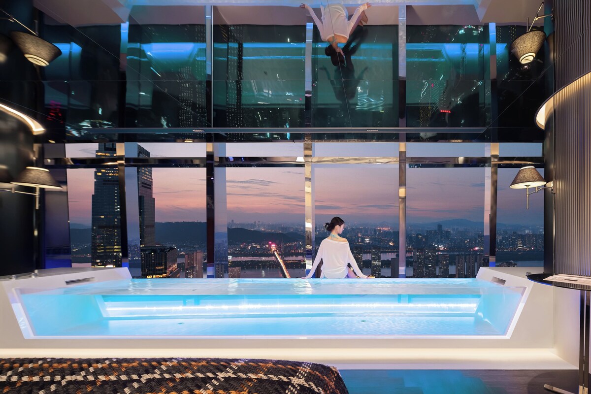 A modern hotel room with a city view, a glass-enclosed balcony, and a pool area.