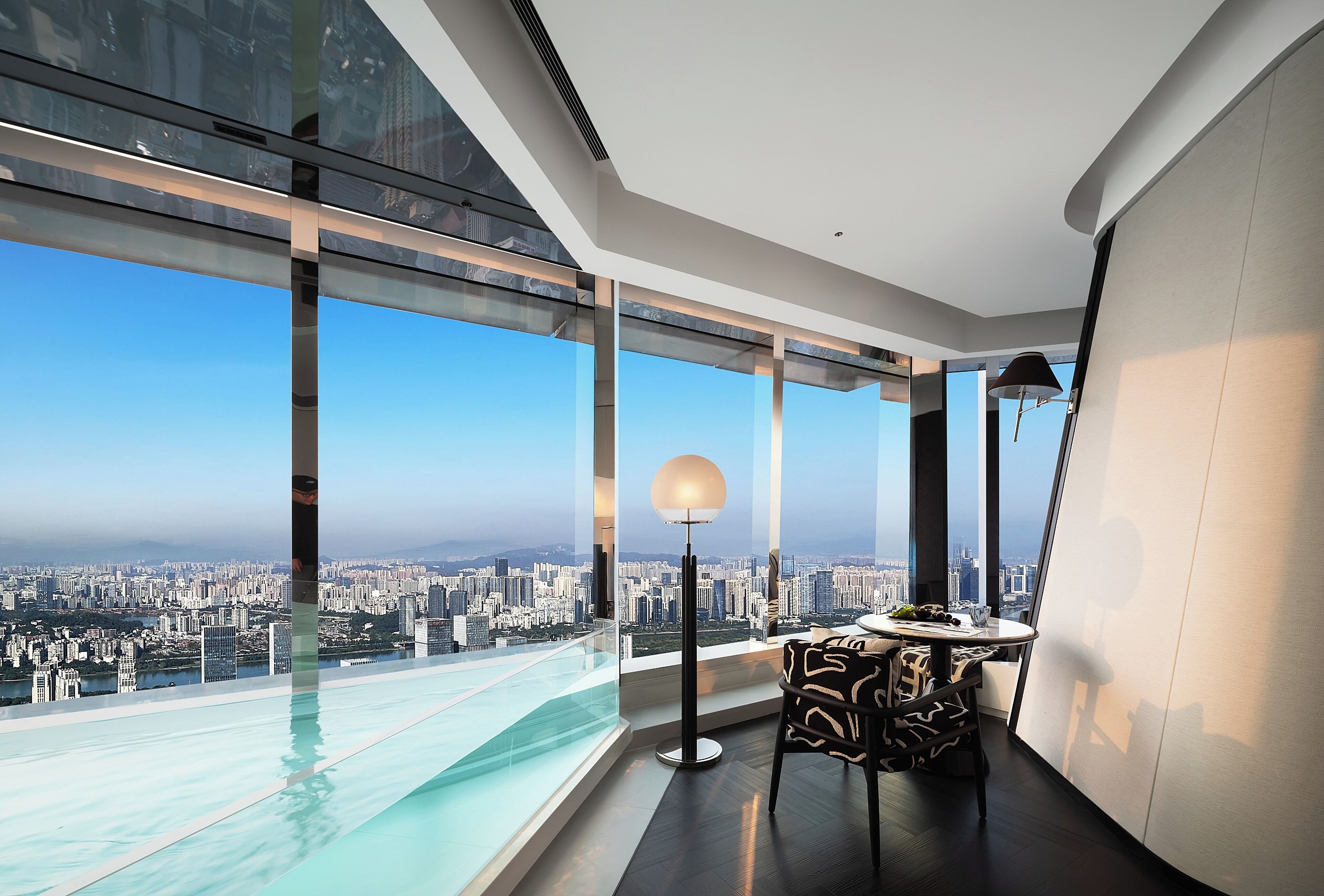 A modern room with a panoramic city view, a glass infinity pool, a round table with a lamp, and a patterned chair.