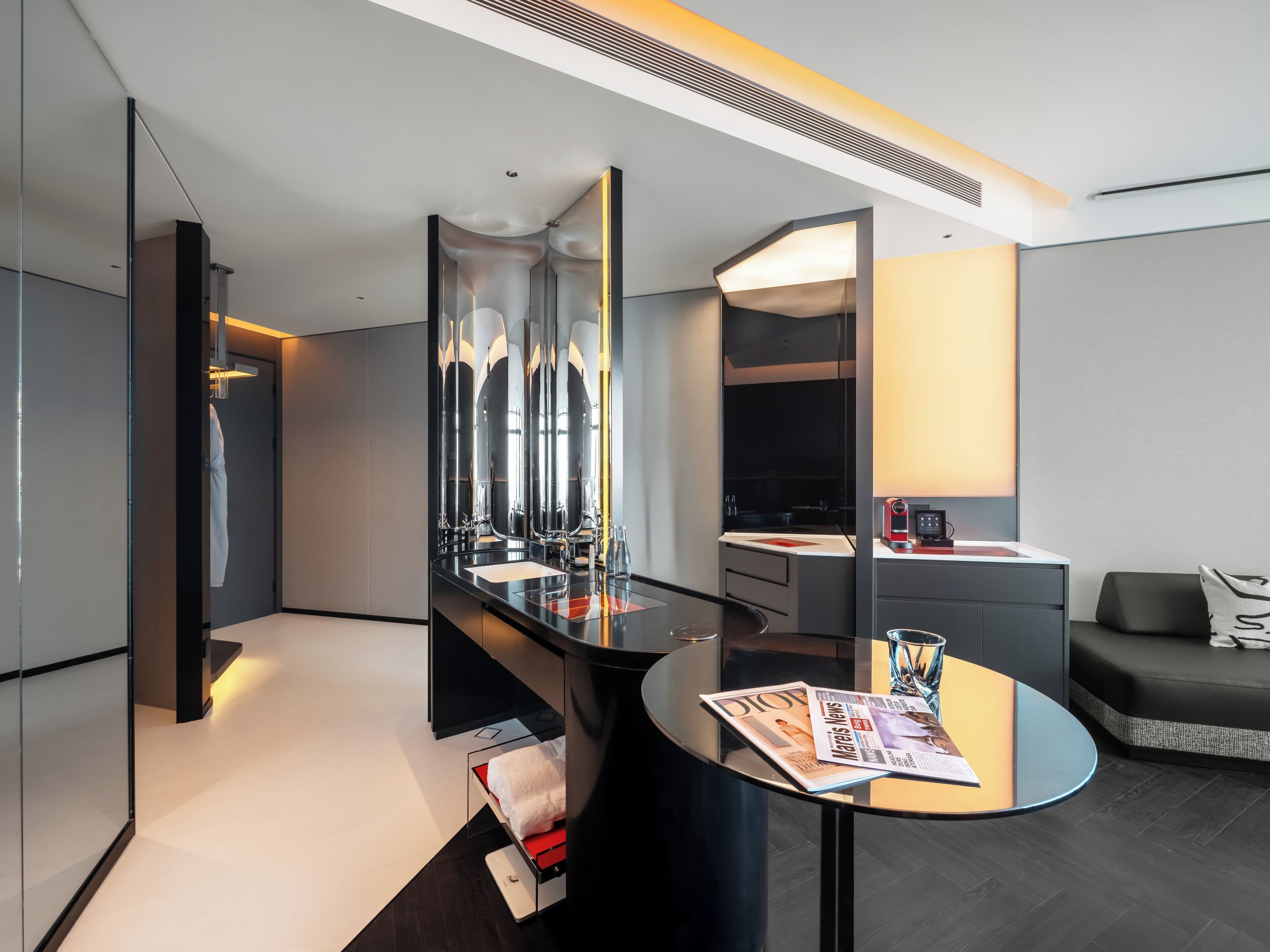 A modern hotel room with a large mirror, a round table with a magazine, a sofa, and a kitchenette area.