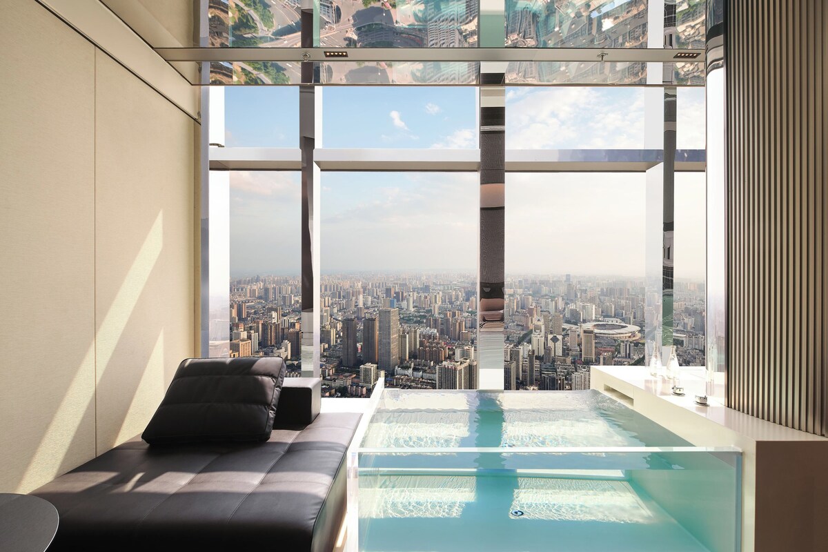A modern room with a large window overlooking a cityscape, a glass-bottomed pool, and a black sofa.