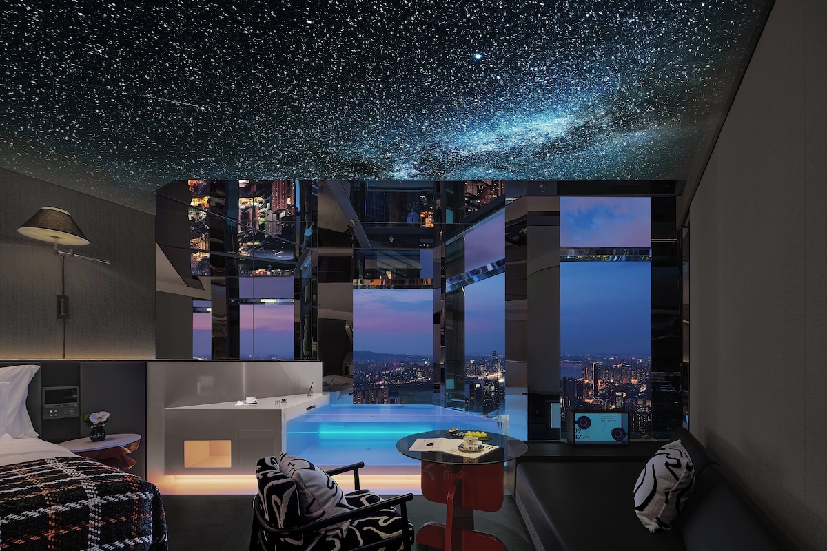 A modern hotel room with a starry sky ceiling, a city view, a bed, a desk, and a small pool area.