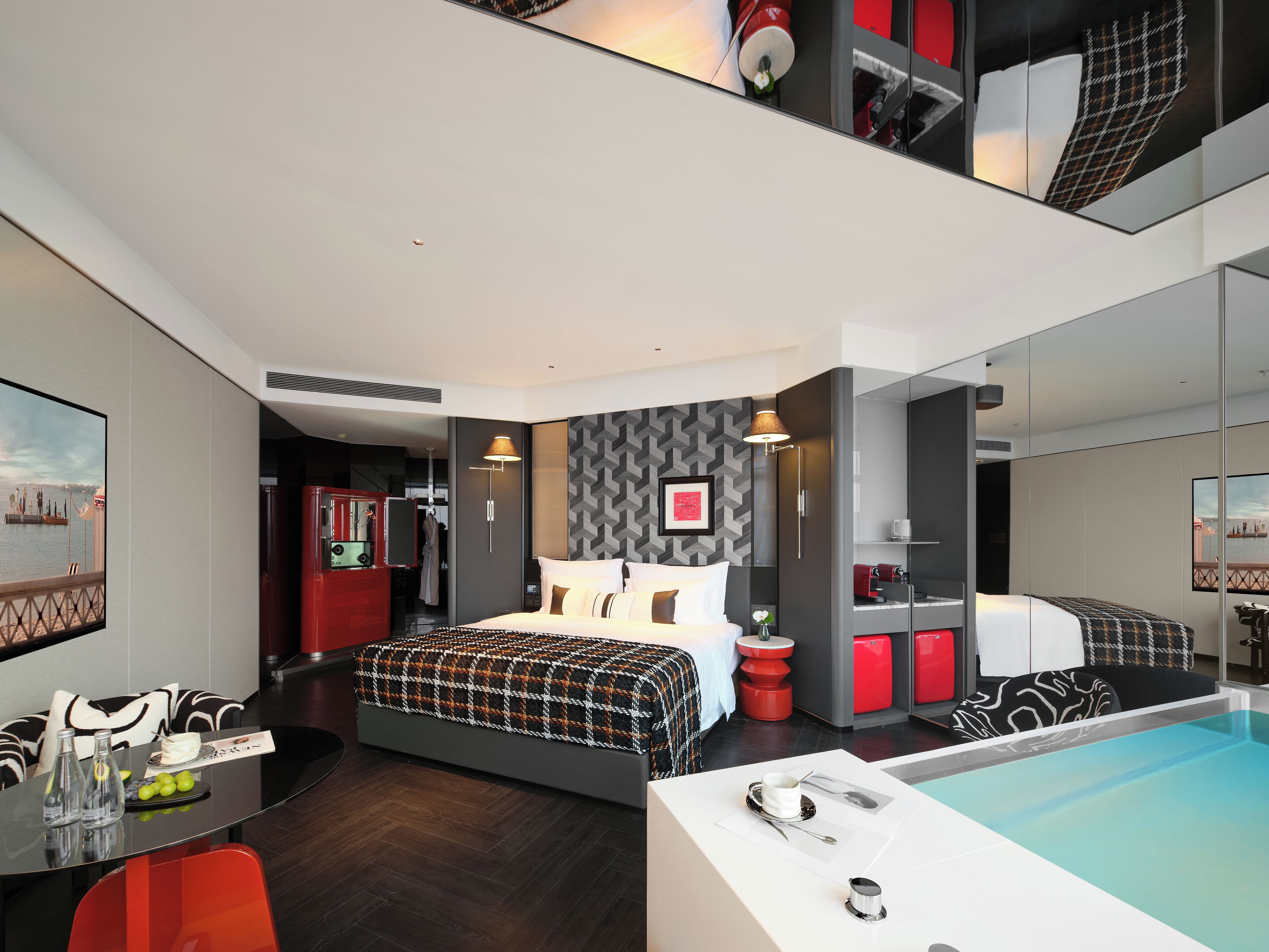 A modern hotel room with a large bed, a small desk, a red chair, a dining table, and a large mirror.