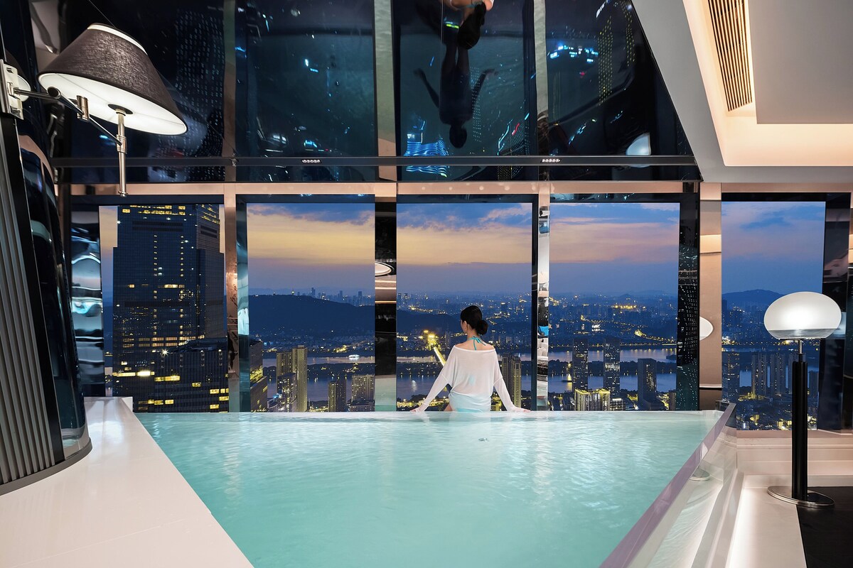 A modern indoor pool with a city view, large windows, and contemporary lighting fixtures.