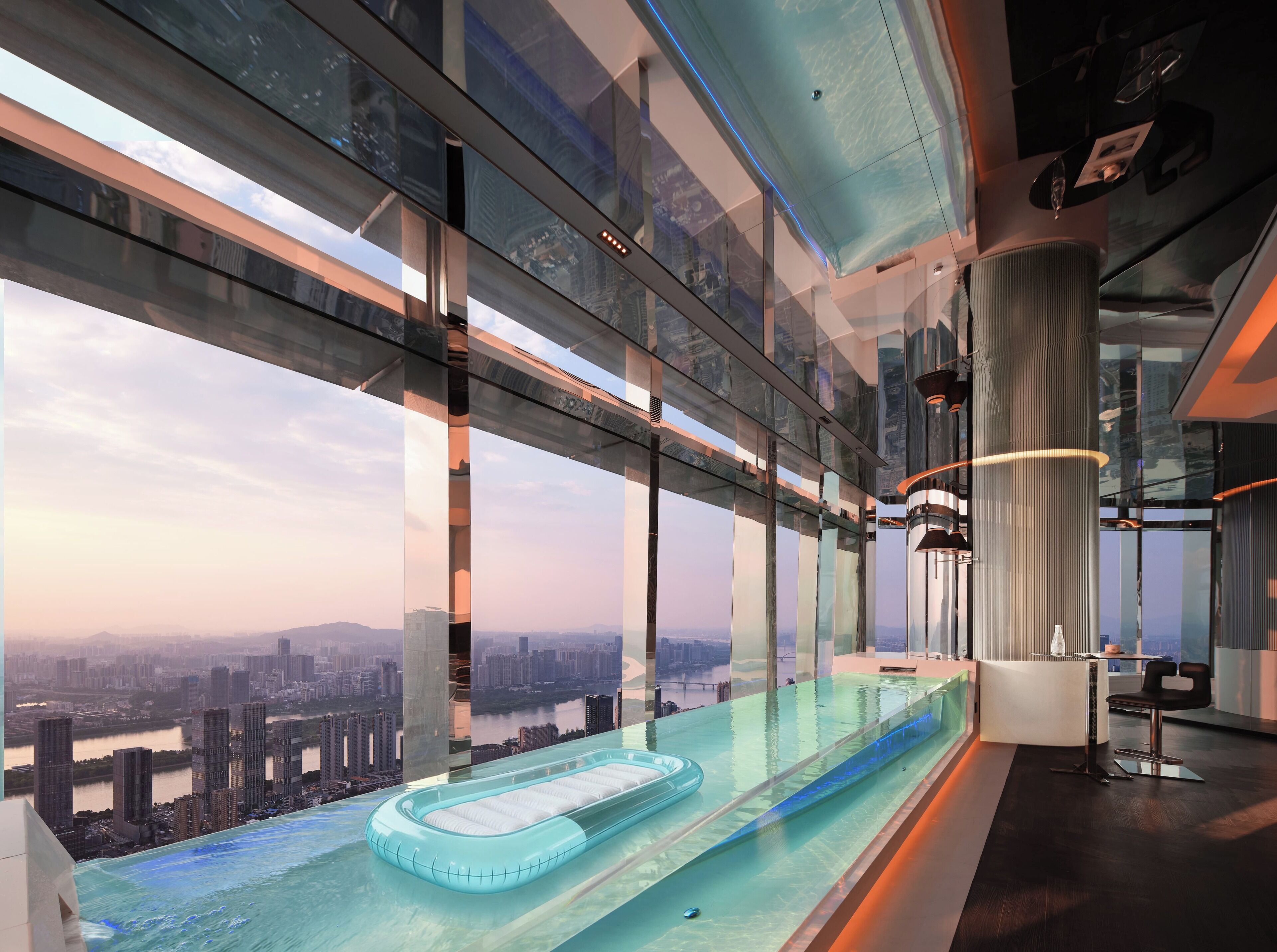 A modern hotel interior with a glass infinity pool, panoramic city views, and contemporary furnishings.