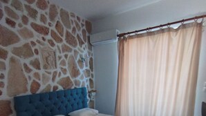 Ein Schlafzimmer mit einer Steinwand, einem blauen Kopfteil, einem Bett mit weißer Bettwäsche und einem Fenster mit beigen Vorhängen.