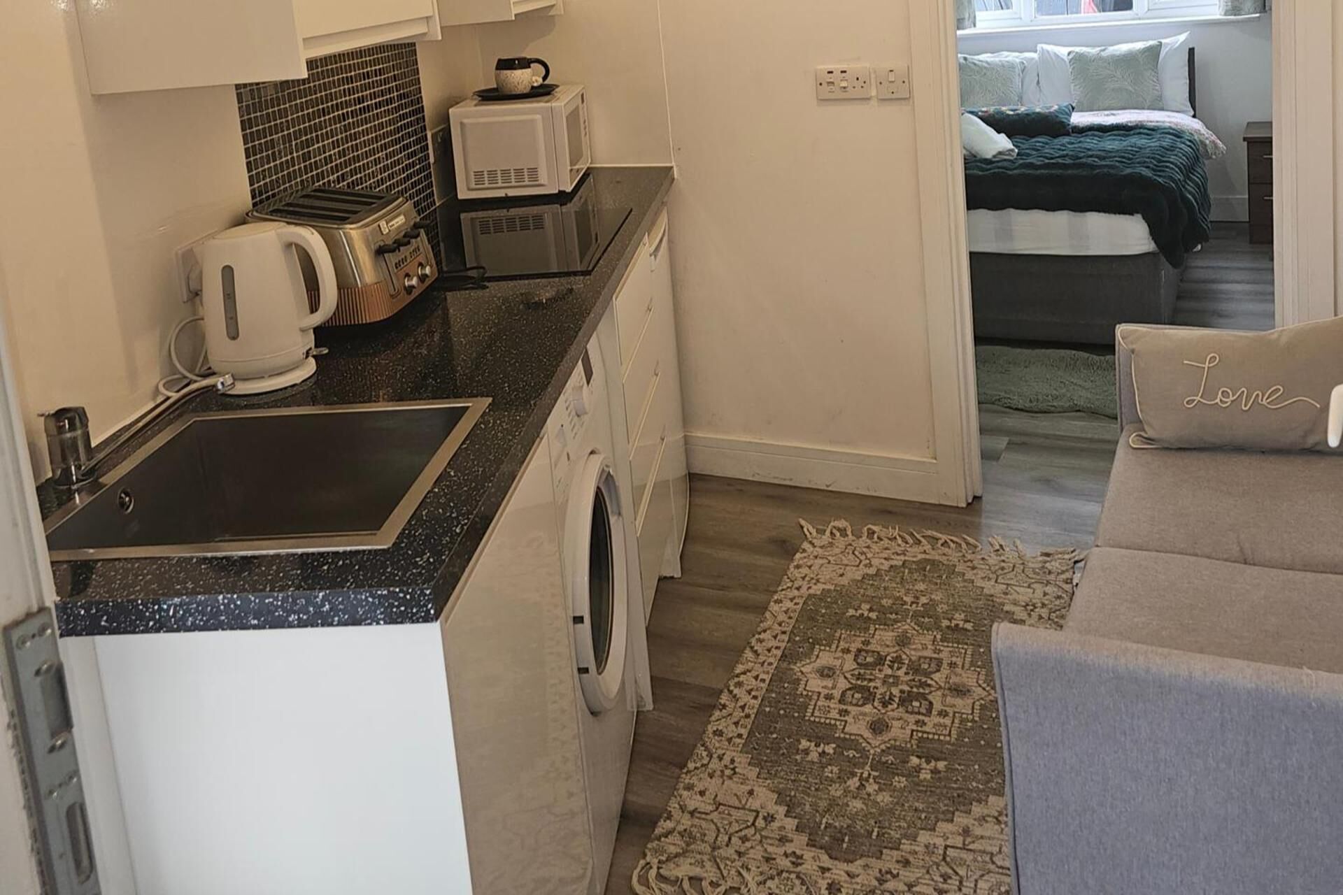 Una cucina compatta con un tostapane, un bollitore e un forno a microonde, un piccolo lavandino e un tappeto che conduce a una camera da letto con un letto e un comodino.
