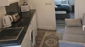 Una kitchenette compacta con microondas, tostadora y tetera, lavadora, una cama con una colcha verde y un sofá con un cojín que dice 'Love'.