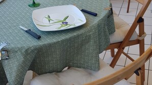 A dining table with a green tablecloth, two square plates with a leaf design, two green wine glasses, a blue pen, and a knife on the table. There are wooden chairs with white cushions around the table. - T2 exceptional marina view (Arcachon)