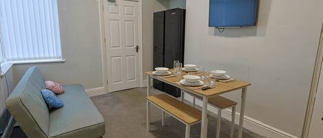 A compact living space with a kitchenette, a dining table set for four, a sofa with cushions, and a television mounted on the wall.