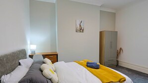 A neatly made bed with white and gray bedding, a yellow blanket, and a fluffy pillow. There is a gray wardrobe and a small table with a lamp in the room.