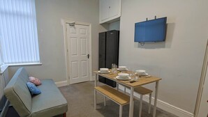 A compact living space with a kitchenette, a dining table set for four, a sofa with cushions, and a television mounted on the wall.