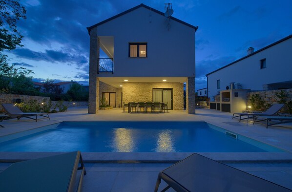 A two-story house with a swimming pool, outdoor seating, and a stone facade.