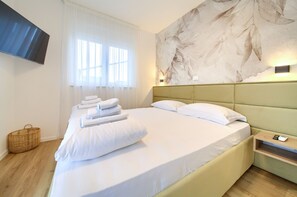A neatly made bed with white linens and towels, a green headboard, a wall-mounted TV, and a wooden floor.