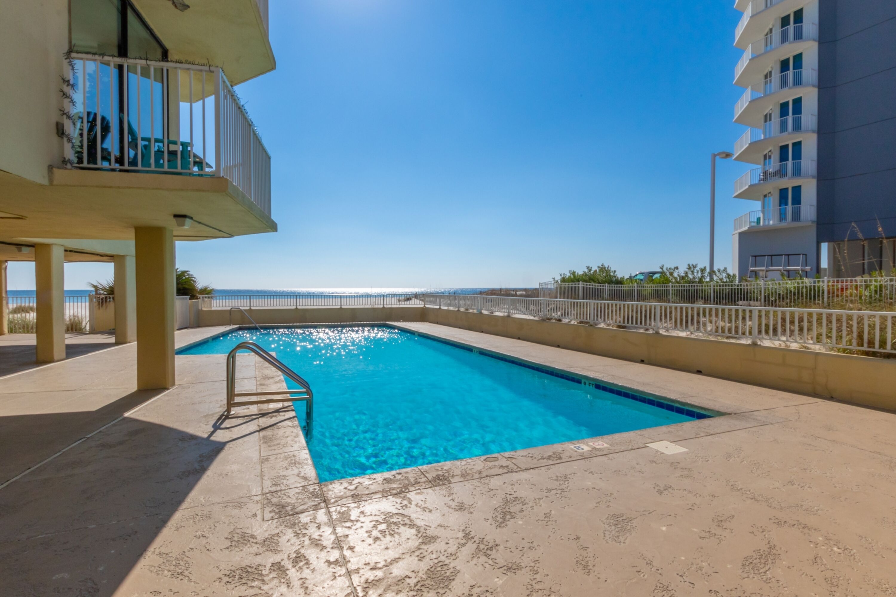 A swimming pool with a clear blue water, surrounded by a concrete deck and a white fence.