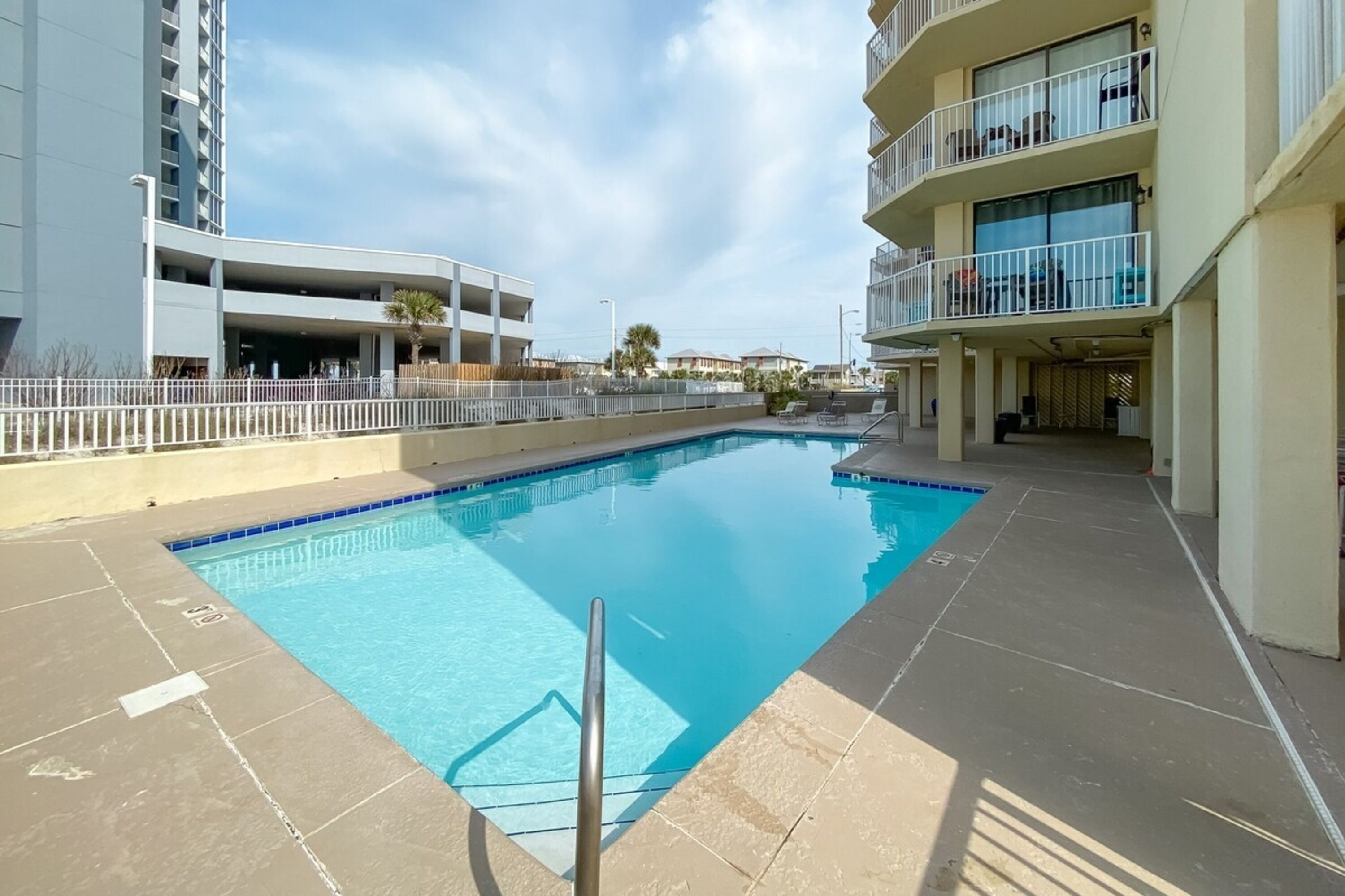 A swimming pool with a clear blue water surface, surrounded by a white fence and adjacent to a multi-story building with balconies.