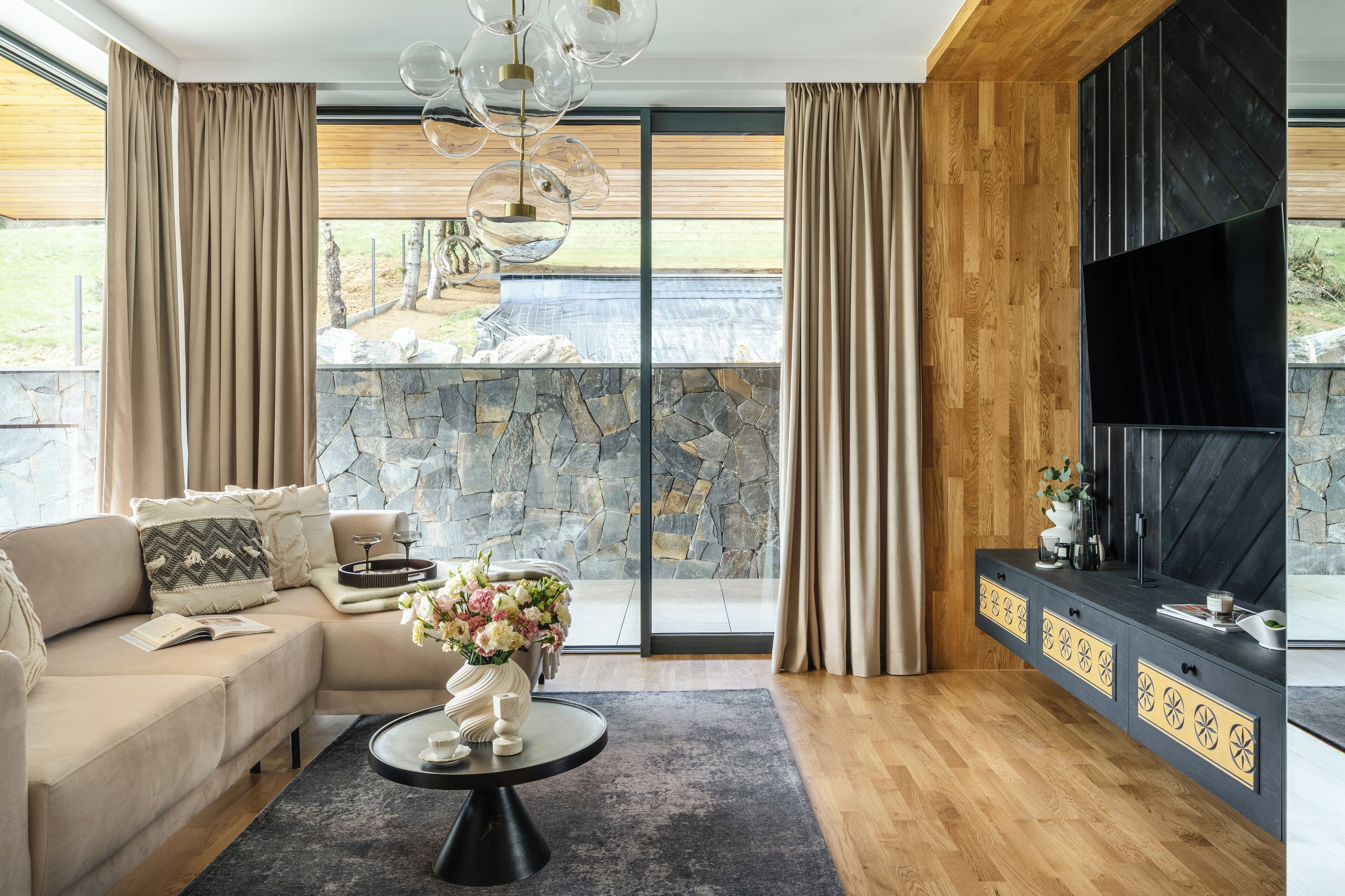 A modern living room with a stone fireplace, a sofa, a coffee table with a vase of flowers, and a large TV on a wooden wall.