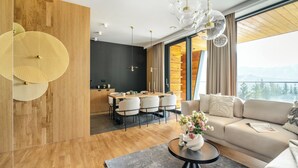 A modern living room with a wooden floor, a sofa, a dining area with a table and chairs, and a large window with a view of mountains. - Salamandra Luxury Villas with Jacuzzi (Kościelisko)