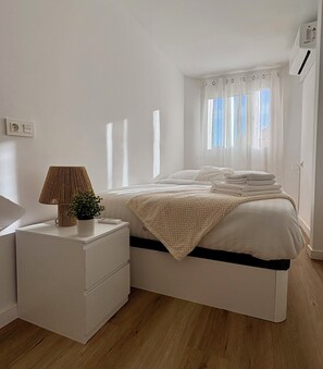 Een minimalistisch ingerichte slaapkamer met een bed, nachtkastje en een raam met gordijnen.