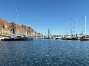 Een jachthaven met verschillende zeilboten die aangemeerd liggen, een rotsachtige heuvel op de achtergrond en rustig water.