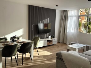 Een moderne woonkamer met een eethoek, een flatscreen-tv aan de wand, een bank en een raam met gordijnen.