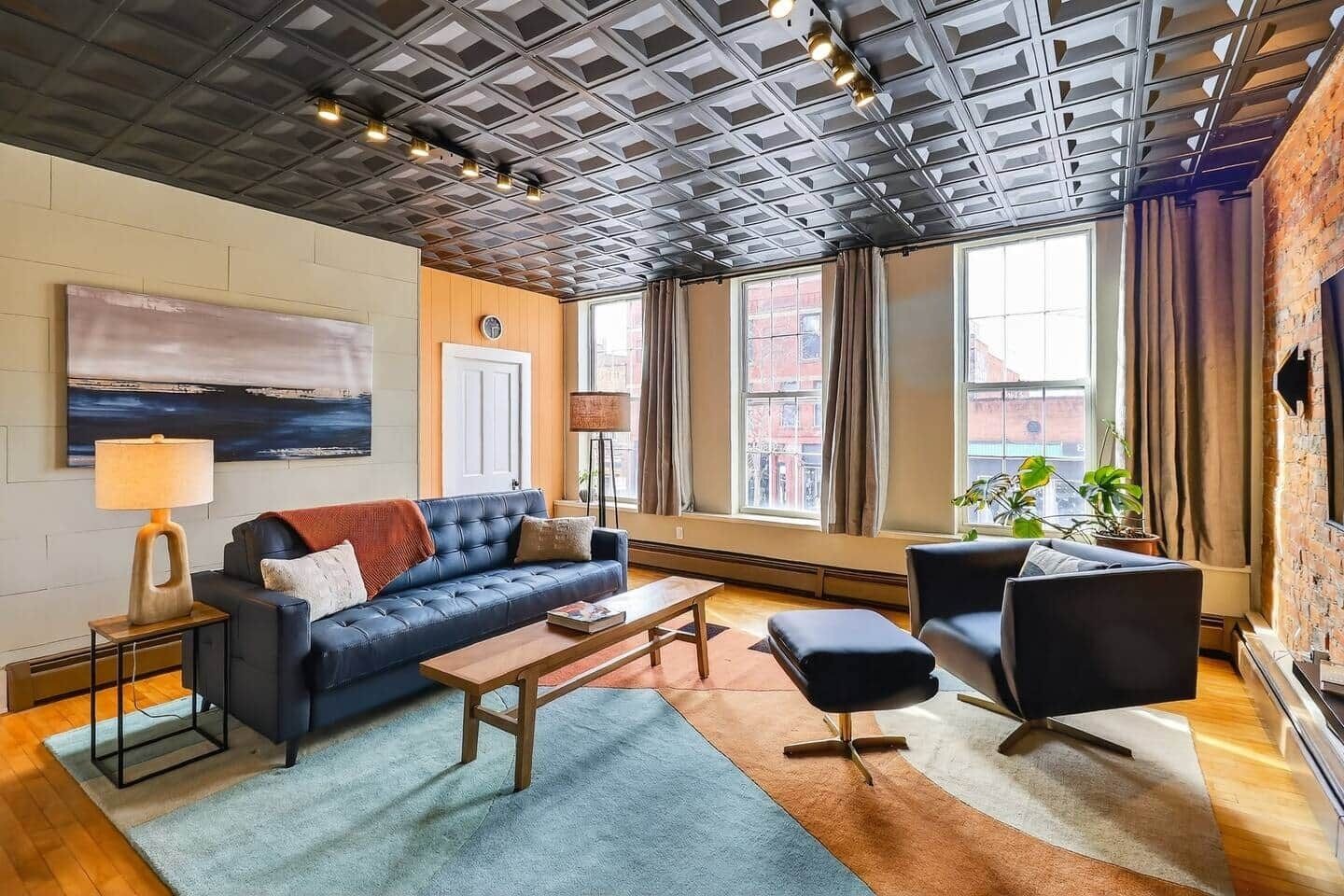 Un salon moderne avec un canapé bleu, une table basse en bois et un fauteuil noir. Il y a une grande fenêtre avec des rideaux, un tableau au mur et une lampe sur une table d’appoint.