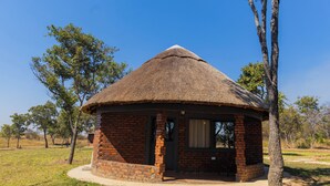 A thatched-roof structure with brick walls, a door, and a window, surrounded by grass and trees. - Spring Resort  (Hurungwe)