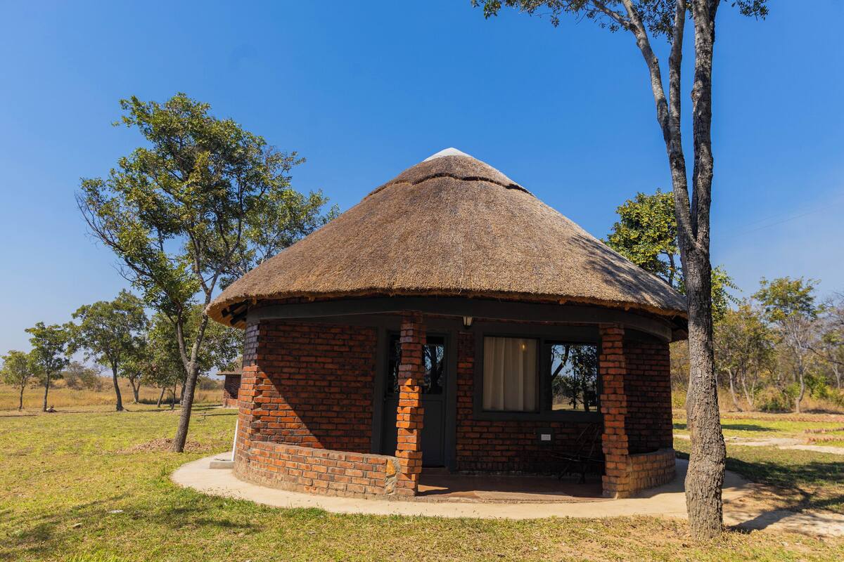 A thatched-roof structure with brick walls, a door, and a window, surrounded by grass and trees.