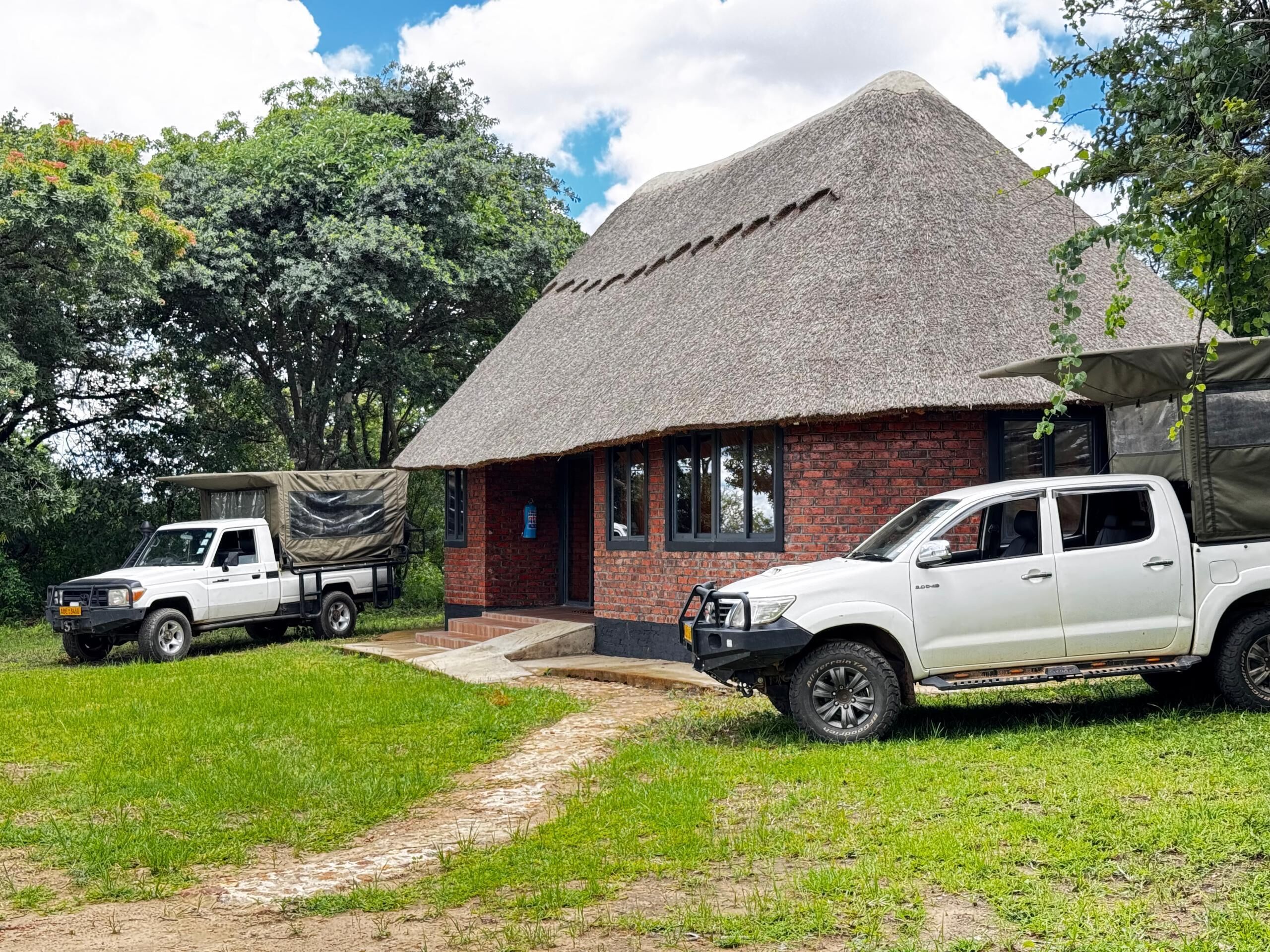 A thatched-roof building with a brick exterior, a covered outdoor seating area, and two white pickup trucks parked in front.