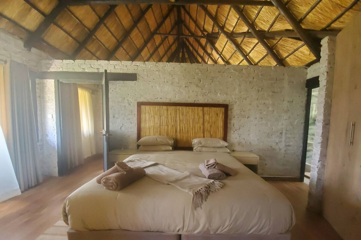 A spacious bedroom with a large bed, a wooden headboard, and a thatched roof ceiling.