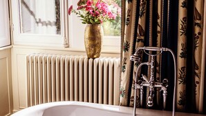 Une salle de bain avec une baignoire sur pieds, un radiateur, une fenêtre avec des rideaux et un vase de fleurs posé sur le rebord de la fenêtre.