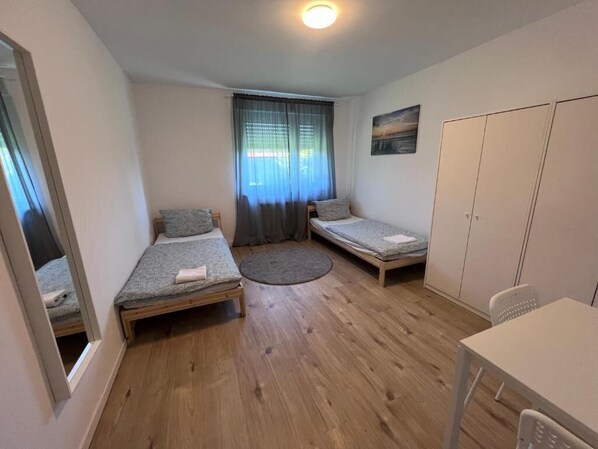 Una habitación pequeña y bien iluminada con dos camas individuales, un tapete redondo, una mesa y un armario.