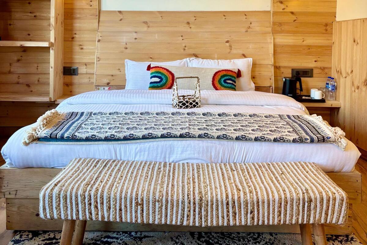 A wooden-paneled bedroom with a large bed, two rainbow-patterned pillows, a decorative wall piece, and a bench.