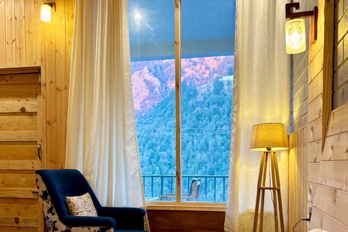 A cozy room with a bed, a chair, a small table, and a window with a view of a forested mountain.