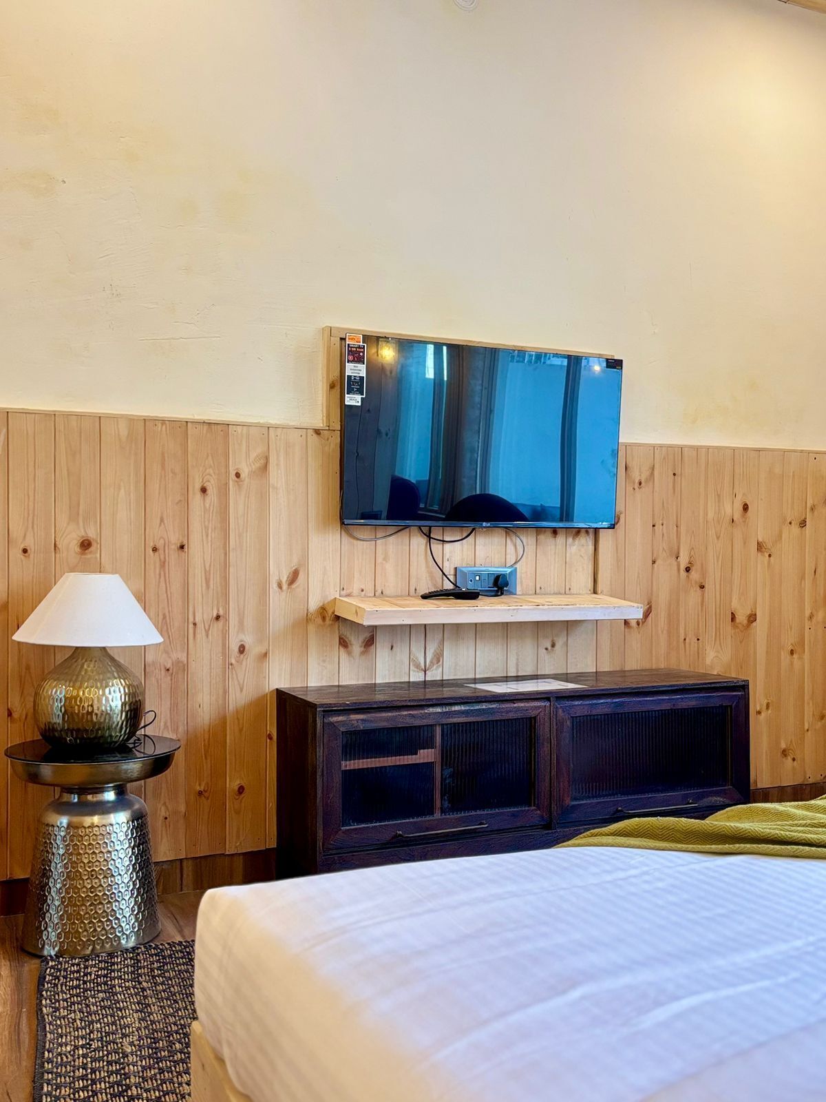 A hotel room with a bed, a TV mounted on a wooden wall, a lamp, and a small wooden cabinet.