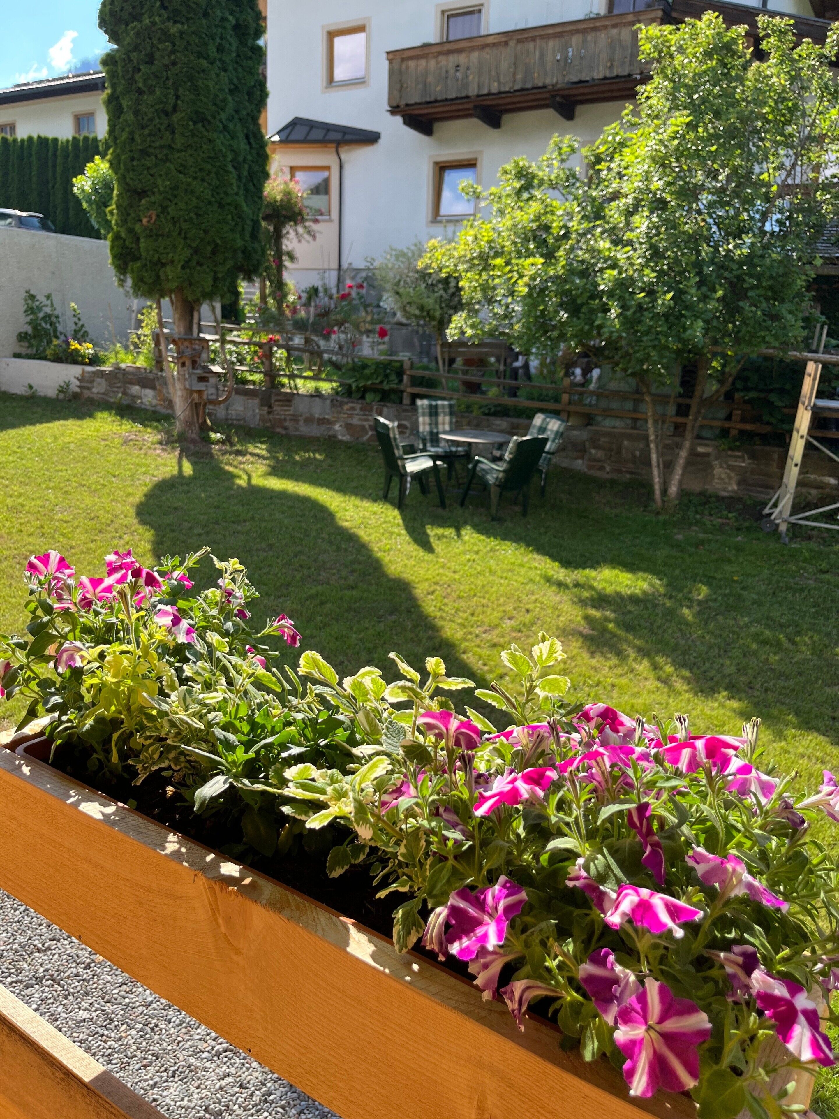 Un jardin avec un parterre en bois rempli de fleurs roses, un mur de pierre et un bâtiment avec un balcon en arrière-plan.