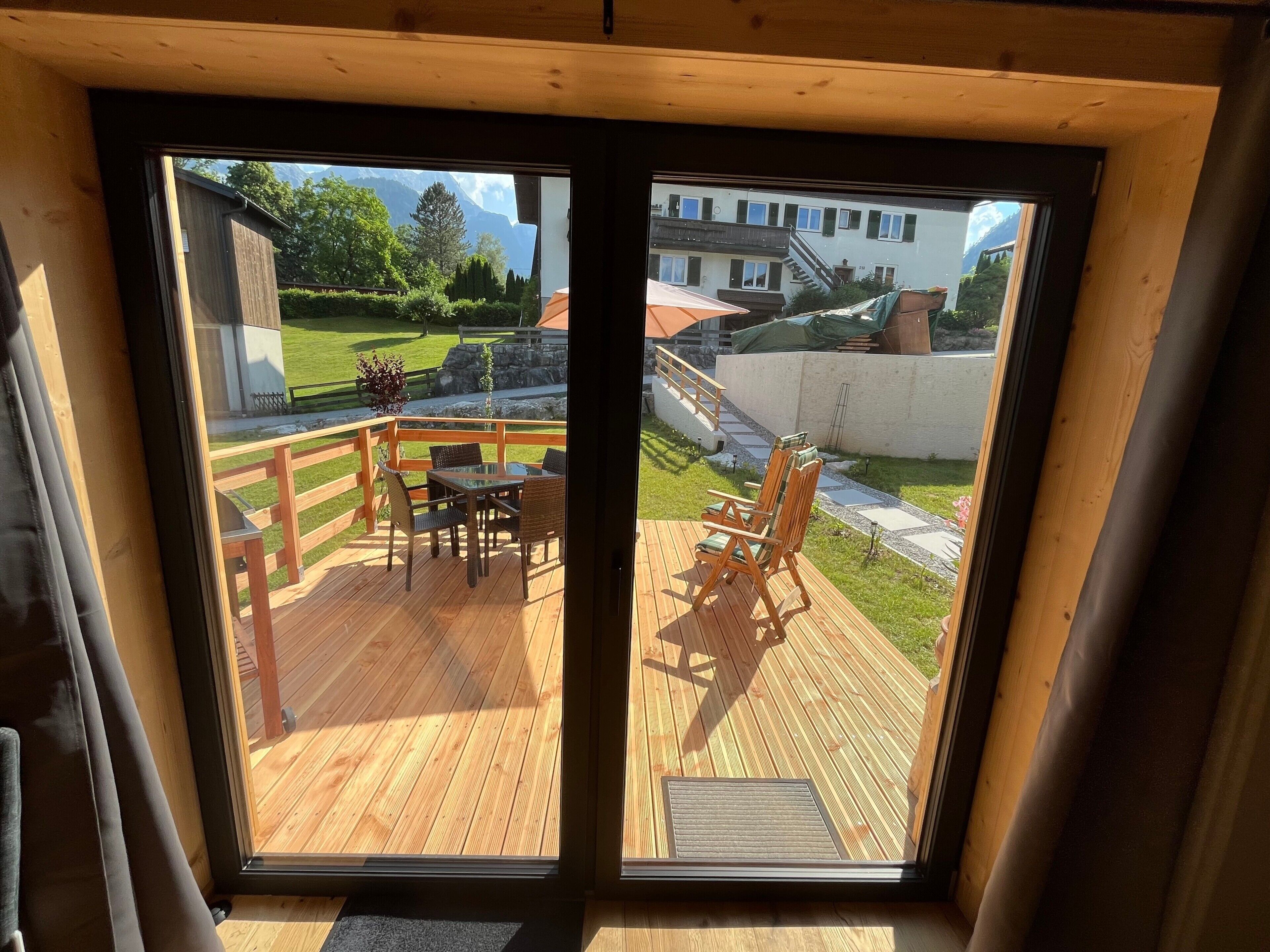A wooden deck with outdoor furniture, a view of a garden, and a building in the background.