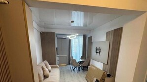 Een moderne woonkamer met een bank, een eettafel en stoelen. Er is een groot raam met gordijnen, een plafondlamp en een aan de muur bevestigde televisie.
