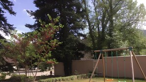 Une aire de jeux avec des balançoires et une structure d’escalade, entourée d’arbres et d’un mur.