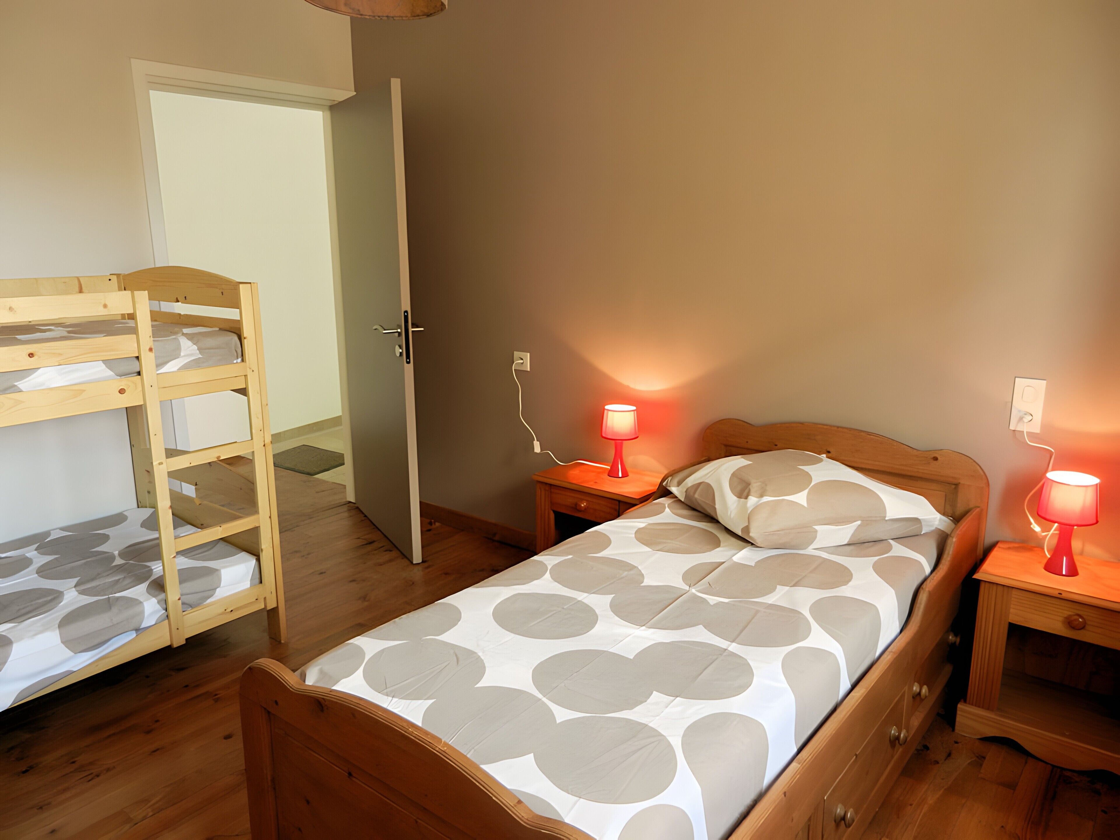 Une chambre avec un lit superposé, un lit simple avec une literie à motifs, deux tables de chevet avec des lampes et une porte donnant sur une autre pièce.