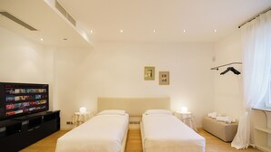 Habitación de hotel con dos camas individuales, televisor sobre un soporte, mesita de noche, lámpara de noche, banco con toallas y ventana con cortinas.