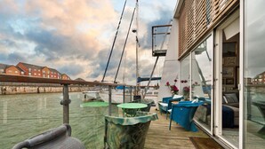 Une terrasse dotée d’une rambarde en verre, d’une table et de chaises, offrant une vue sur une étendue d’eau où l’on aperçoit des bateaux et des bâtiments au loin.