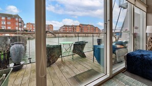 Un balcon donnant sur une rivière, une terrasse en bois et un lit recouvert d’une couverture bleue.