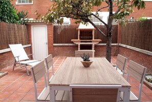 Eine Terrasse mit Holztisch und -stühlen, einem kleinen Topfpflanzen und einem Grill aus Ziegelsteinen.