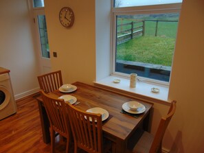 Un coin repas avec une table en bois, quatre chaises, une horloge au mur et une fenêtre donnant sur un champ verdoyant.