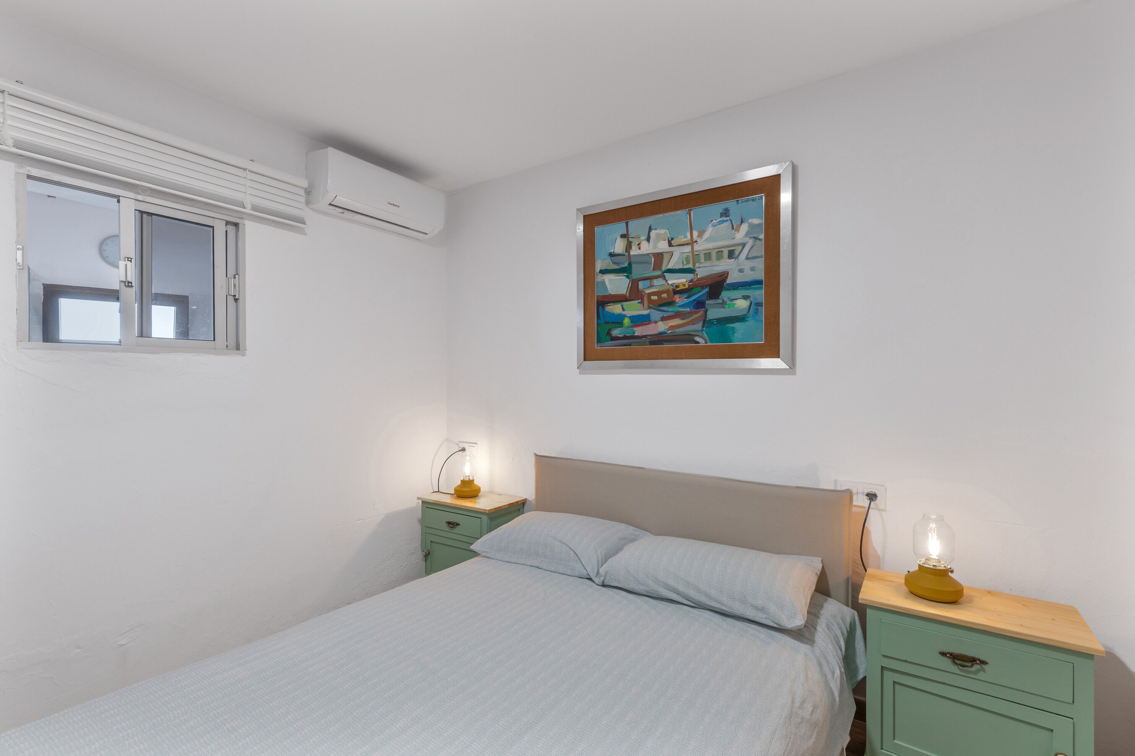 Une chambre à coucher comprenant un lit, deux tables de chevet, une commode, un climatiseur fixé au mur, une fenêtre et un tableau.