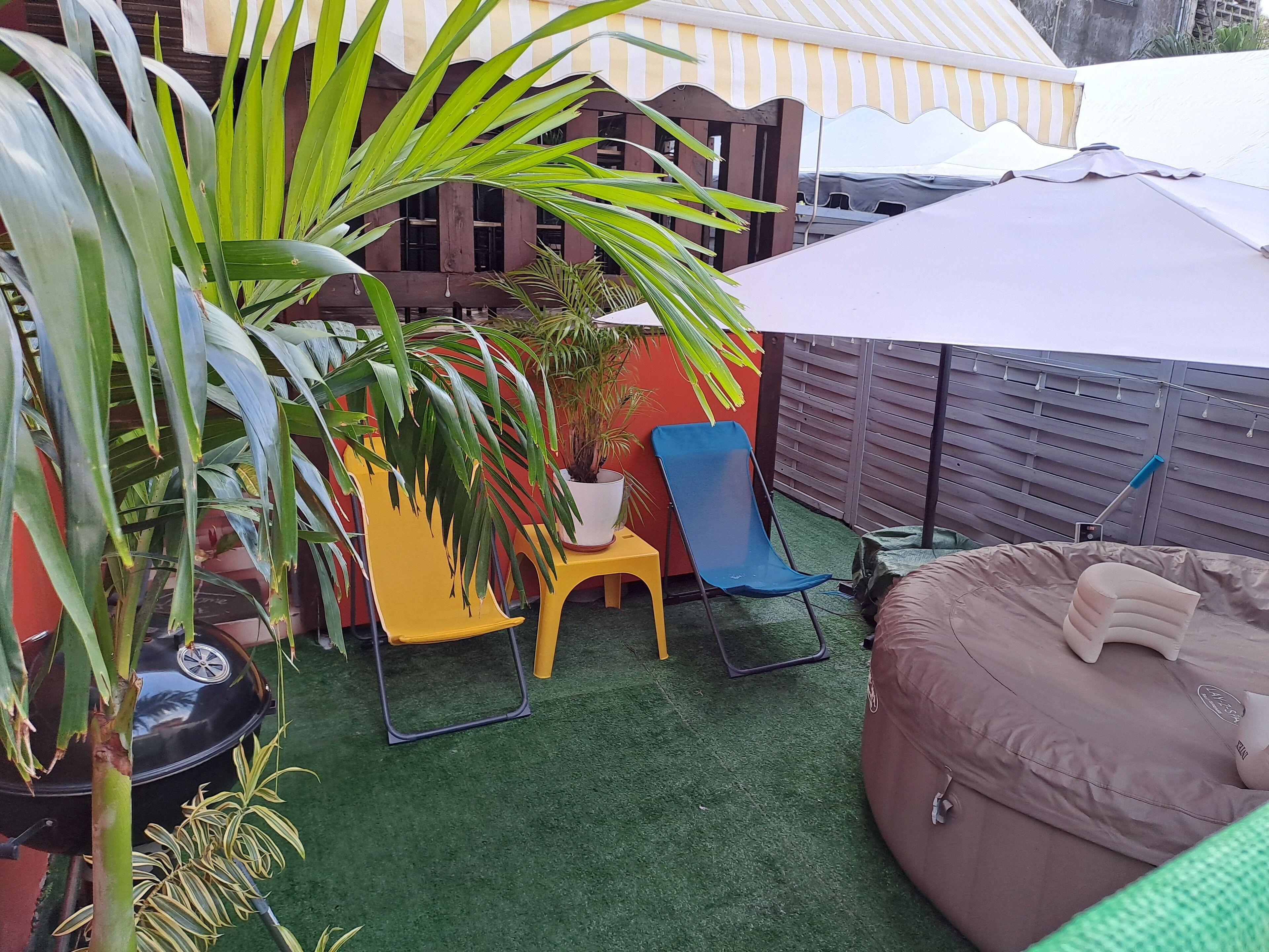 Un espace patio avec une pelouse artificielle verte, une chaise jaune, une chaise bleue, un grand parasol et un fauteuil en forme de sac.